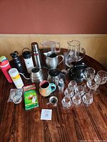 Full set on a round wooden table against a red wall. Various drinkware including water bottles, thermal bottle, glass pitchers, wine glasses, and a black wine aerator.
