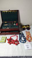 Wood jewelry box with compartments for earrings, rings, and necklaces. Several pieces of costume jewelry laid out in front and beside the box including necklaces and two pairs of sunglasses.