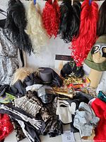 Photo showing multiple wigs hanging in black, white, red, and blonde shades, assorted masks including paper mache and animal style masks, miscellaneous costume accessories and fabric pieces on a flat surface.