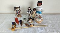 Collection of indigenous-themed dolls, feathers, pipe, beaded necklace, and birch bark basket with fur lining on white fabric.