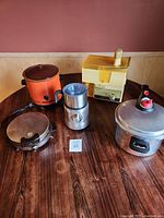 Full view of five kitchen appliances including orange vintage crockpot, silver juicer, yellow and cream pressure cooker, brown waffle iron, and small silver foamer on wooden table.