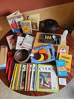 Top view of kitchen collection on round wooden table showing vintage cookbooks, cast iron pot, utensils, coasters, butcher block, cheese board
