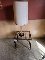 Full view of tall lamp with beige fabric cylinder shade sitting on gold bamboo glass top end table against wall