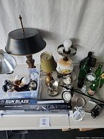 Photo showing multiple lamps including a pineapple base lamp, floral glass lamp base missing shade, a black metal desk lamp, and various glass bottles on a table.
