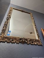 Full view of large rectangular gold framed wall mirror mounted on a dark gray wall, showing the ornate decorative frame and reflective glass.