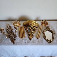 Full display of eight decorative gold items including mirror, sconces, ceramic candleholders, hanging shelf, and gold taper candles arranged on white cloth