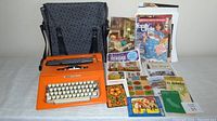 Photo of orange Olivetti Lettera 25 typewriter with grey patterned carrying case and various vintage paper ephemera including magazines and maps spread on table