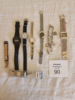 Lot contains seven watches and a silver-tone heart charm bracelet laid out on a light cloth background, showing variety of watch styles and straps.