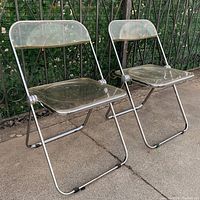 Two clear acrylic and chrome folding chairs set up outside on concrete with fence background, visible wear on acrylic seats and backs.