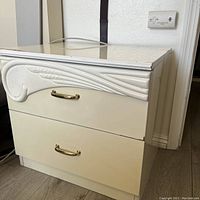 Front view of cream lacquer nightstand showing two drawers with gold-tone handles and decorative carved detail on the top drawer