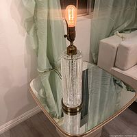 Full view of the lamp on a reflective surface with light bulb illuminated, showing the crystal cut column and brass base.