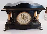 Front view of antique Seth Thomas mantle clock with black and faux marble finish, showing ornate columns, brass feet, and clock face.