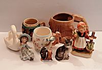Mixed lot showing all 8 pieces: 2 souvenir mugs, a German hunting stein, child's whistle cup, cat figurine, swan figurine, and two young girl figurines.