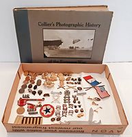 Photo showing the vintage Collier's Photographic History book with accompanying assorted vintage military pins, buttons, patches, mini flags, and a white multi-tool knife laid out in a cardboard tray.