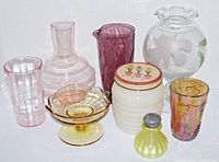 Group image of seven vintage glassware pieces. Pink glass carafe and tumbler, purple cocktail mixer, large clear orchid-decorated glass, ribbed white jar with tulip lid, yellow ribbed perfume bottle, iridescent floral tumbler visible.