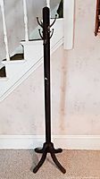 Full view of the tall, dark wood coat rack standing in front of a staircase, showing the four metal hooks and the four-legged base.