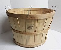 Front angled view of wooden bushel basket showing metal handles and manufacturer marking on wooden slat.