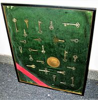 Full view of the 24-piece antique key pendulum collection framed behind glass with green velvet background and a brass medallion center