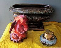 Three art pottery pieces arranged on yellow cloth: flambe dragon figurine, ornate Beauceware vase, blue mountain pottery lighter