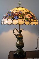 Front view of the lamp showing the stained glass floral shade and bronzed metal amphora base on a black marble platform.