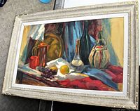 Framed oil painting showing a colorful still life scene with bottles, fruits and drapery, artist signed H. A. Ellam at bottom left.