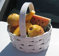 Photo showing the white porcelain weave basket filled with carved alabaster fruit figurines including pineapple, banana, watermelon, peach.
