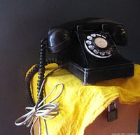 Black Northern Electric rotary dial telephone angled view showing coiled handset cord and plug-in cord coiled in front, sitting on yellow cloth against dark background.