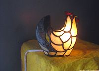 Front and side views of a stained glass chicken-shaped table lamp illuminated, showing cream colored panels with red comb and eye, and dark tail feathers.