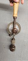 Close-up of the wooden handle, metal gear wheel with teeth, and crank mechanism of the antique hand drill showing wear and patina.