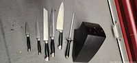 Photo showing 6 knives and honing steel laid out beside the black wood block on a gray floor.
