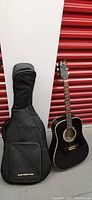 Black Bull Brand acoustic guitar standing next to a black New York Pro soft guitar case with visible wear.