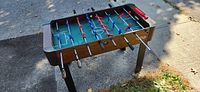 Foosball table visible on concrete ground outdoors with shadows; view of playing field, handles and legs; wood grain finish sides, black handles, blue and red players.