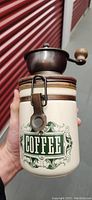 Front view of vintage coffee grinder showing ceramic base with 'COFFEE' label and metal clasp locking mechanism.