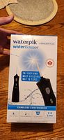 Front of the boxed Waterpik Cordless Plus Water Flosser showing brand, product name, and key features including cordless convenience and 4 tips.