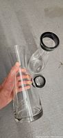  Photo of one glass serving jug held in hand showing clear cylindrical body and black lid with rubber seal detached on table.