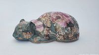 Porcelain sleeping cat figurine lying curled with intricate pink and gold floral painted patterns on teal background.