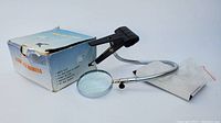 Magnifying glass with flexible metal neck, black plastic clamp, product box and instruction manual laid out on white surface.