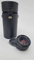 Minolta MD 50mm f/2 lens placed beside its black textured protective cylindrical case with clasp and carrying strap.