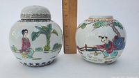 Two round Chinese Famille Rose porcelain jars; one jar with lid featuring a seated woman by banana plants, the other jar without lid showing a child playing under a tree.