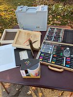 Overall art supplies including wooden case, gray plastic case, trays, and ceramic mug on outdoor table
