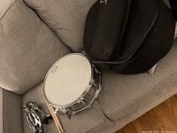 Snare drum, stand, drumsticks, and carrying case arranged on gray couch