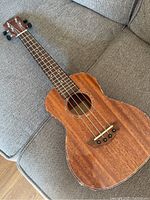 Front and fretboard view of the wooden Mugig ukulele on a grey fabric couch showing natural wood grain and tuning pegs.