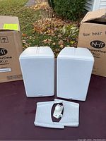 Pair of white NHT SuperOne Xu speakers with original boxes and wall mounting accessories on outdoor table.