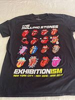 Front view of black T-shirt showing multiple variations of The Rolling Stones tongue and lips logo with event details text at bottom.