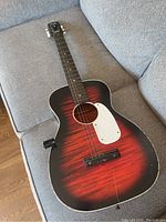Full view of red sunburst Stella parlor guitar on gray couch showing front body, neck, and headstock.