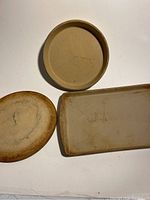 Three stoneware pans arranged on a white background: one round shallow pan, one round deep pan, and one rectangular pan with some visible marks and scratches from use.