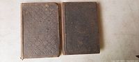 Two very old hardcover books side by side on light surface, showing front covers with textured fabric and aged wear.