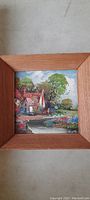Photo of the full painting with wooden frame showing a cottage landscape with lush greenery, flowers, and birds in the sky.