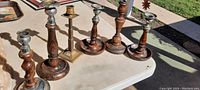 Six antique candlestick holders in brass and wood with varying detailed stems, placed on a white table outdoors.