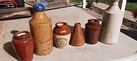 Six early stoneware containers displayed in a row on a table outdoors. Photos show diverse shapes, sizes, and markings.